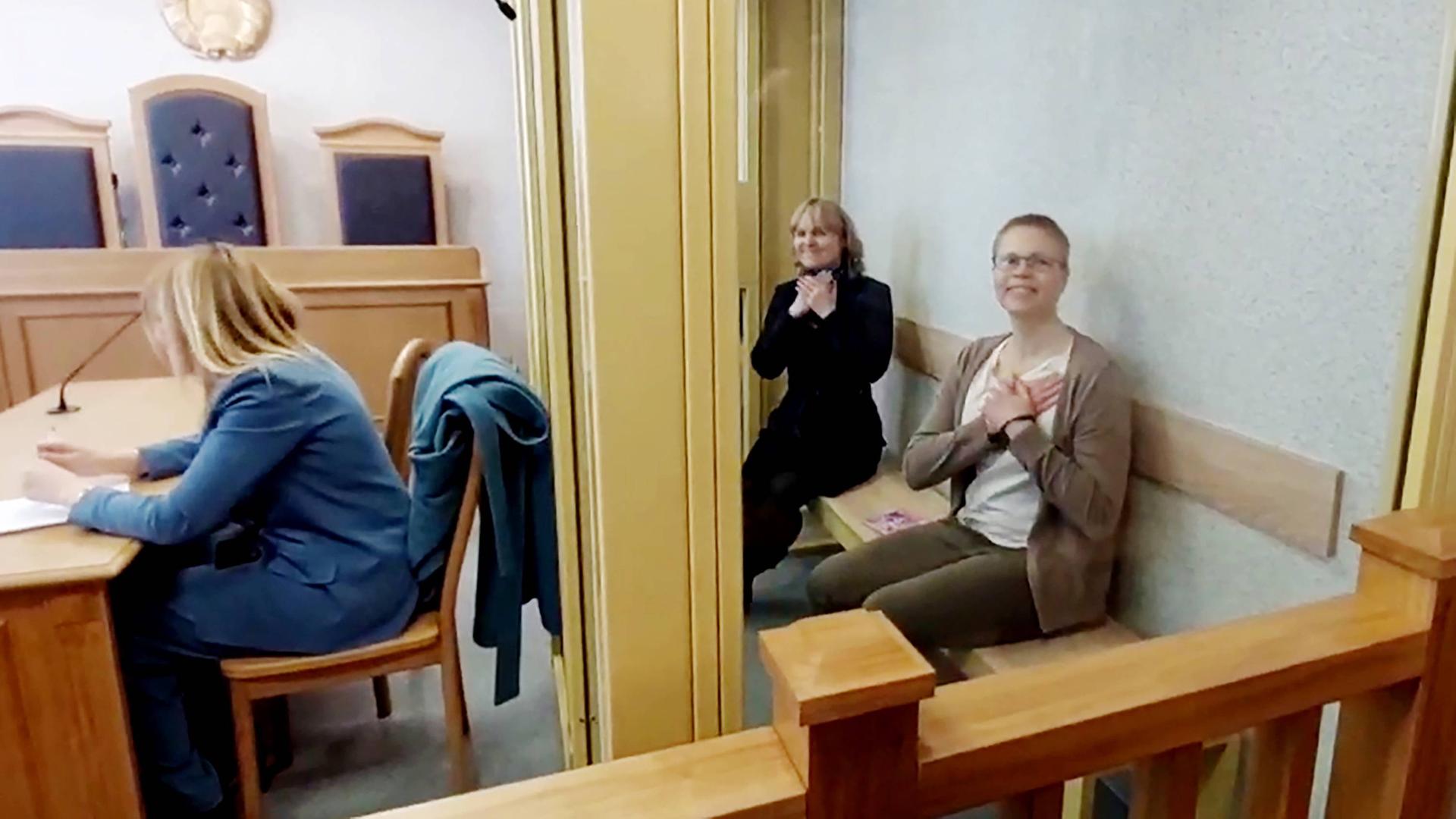 Zwei Frauen sitzen im Gerichtssal in einem Glaskasten und haben die Hände über Kreuz auf die Brust gelegt