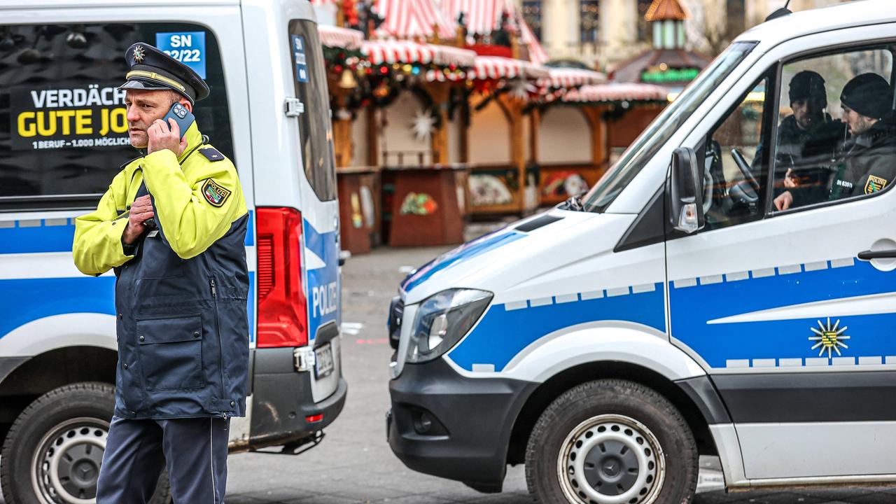 Polizeiwagen sperren die Zufahrt zum Weihnachtsmarkt in Magdeburg. Ein Polizist telefoniert vor zwei Einsatzwagen stehend. Im Hintergrund ist der leere Weihnachtsmarkt zu sehen.