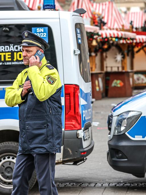 Polizeiwagen sperren die Zufahrt zum Weihnachtsmarkt in Magdeburg. Ein Polizist telefoniert vor zwei Einsatzwagen stehend. Im Hintergrund ist der leere Weihnachtsmarkt zu sehen.