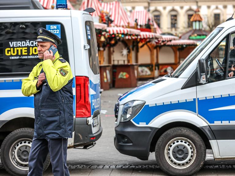 Polizeiwagen sperren die Zufahrt zum Weihnachtsmarkt in Magdeburg. Ein Polizist telefoniert vor zwei Einsatzwagen stehend. Im Hintergrund ist der leere Weihnachtsmarkt zu sehen.