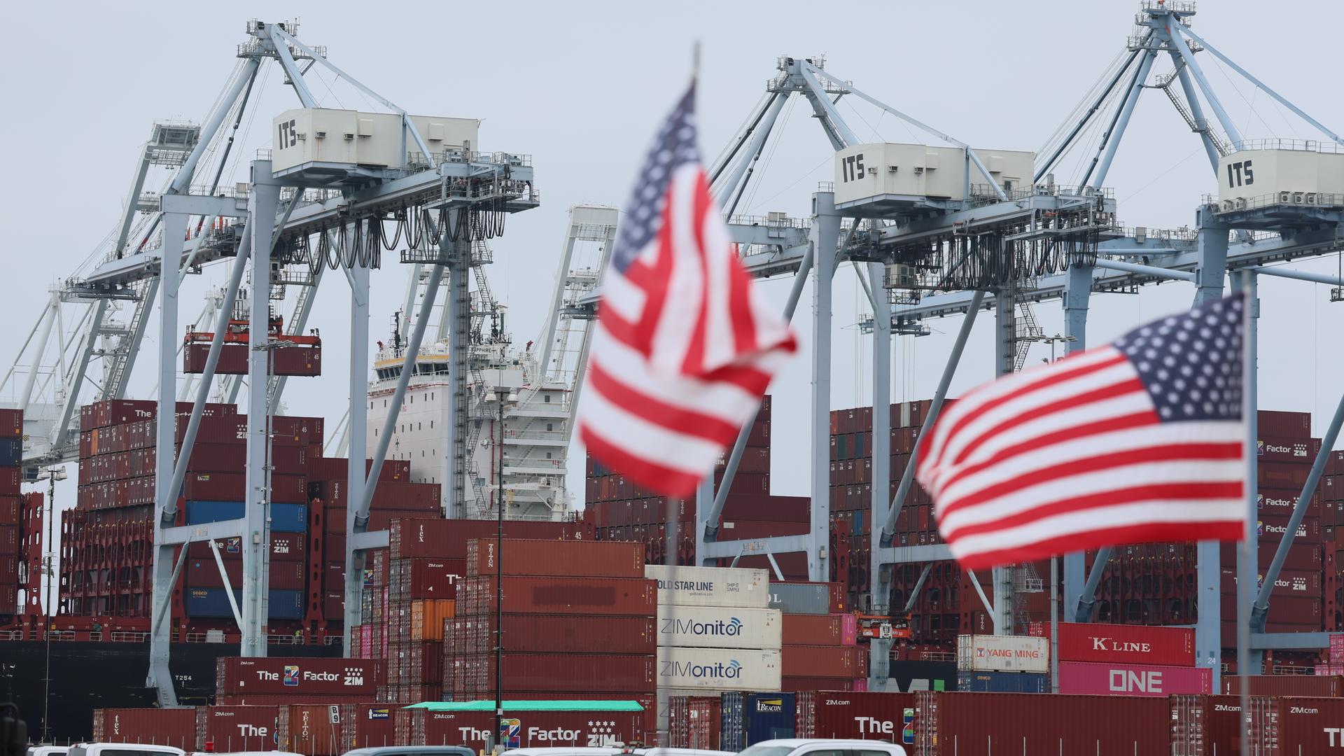 Amerikanische Flaggen vor Containerschiffen und Hafenkränen.
