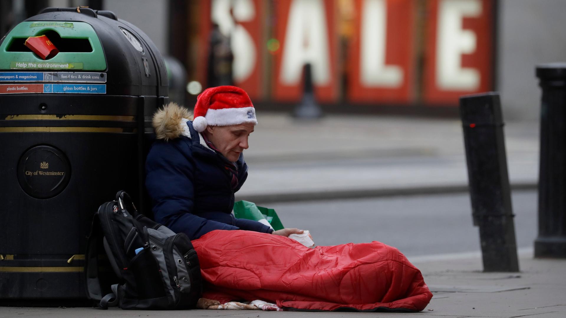 Ein obdachloser Mann mit Nikolausmütze sitzt an einen Mülleimer gelehnt in der Oxford Street in London