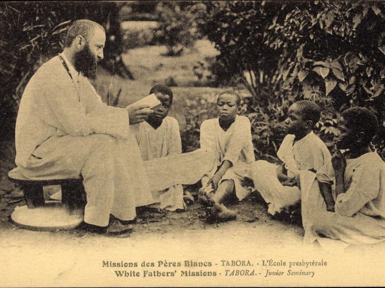 Ein weißer Missionar in Tansania mit afrikanischen Kindern, die vor ihm auf dem Boden sitzen. (1934) 