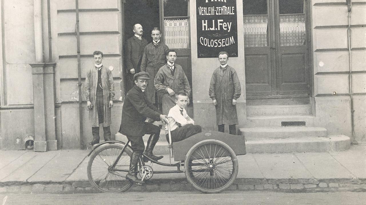 Ein Mann fährt ein Kind in einem Beiwagen vor einem Gebäude mit Filmwerbung. Historische Schwarz-Weiß-Aufnahme um 1920.