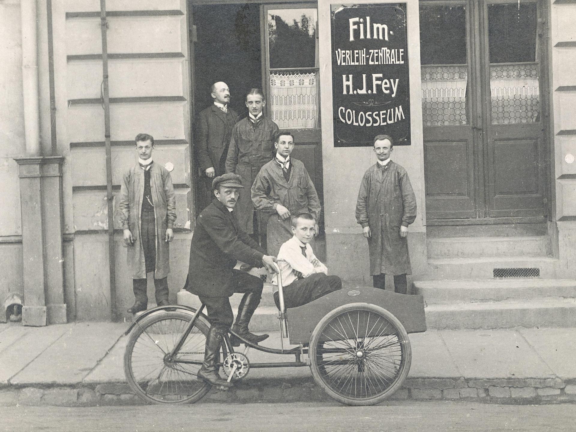 Ein Mann fährt ein Kind in einem Beiwagen vor einem Gebäude mit Filmwerbung. Historische Schwarz-Weiß-Aufnahme um 1920.