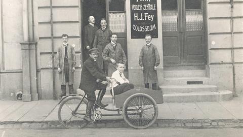 Ein Mann fährt ein Kind in einem Beiwagen vor einem Gebäude mit Filmwerbung. Historische Schwarz-Weiß-Aufnahme um 1920. Ein Mann fährt ein Kind in einem Beiwagen vor einem Gebäude mit Filmwerbung. Historische Schwarz-Weiß-Aufnahme um 1920.