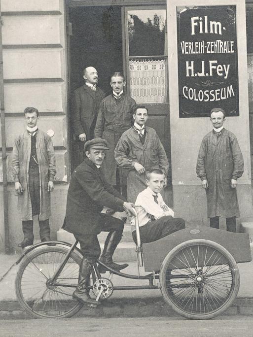 Ein Mann fährt ein Kind in einem Beiwagen vor einem Gebäude mit Filmwerbung. Historische Schwarz-Weiß-Aufnahme um 1920.