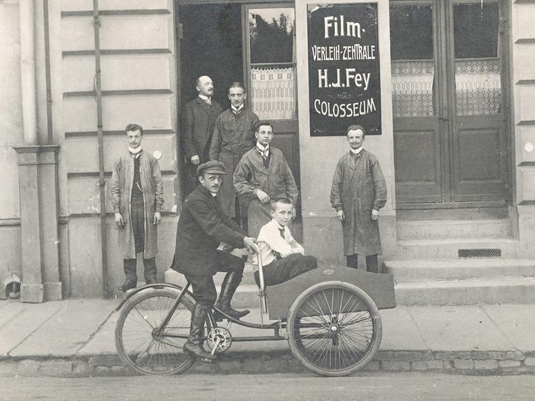 Ein Mann fährt ein Kind in einem Beiwagen vor einem Gebäude mit Filmwerbung. Historische Schwarz-Weiß-Aufnahme um 1920.