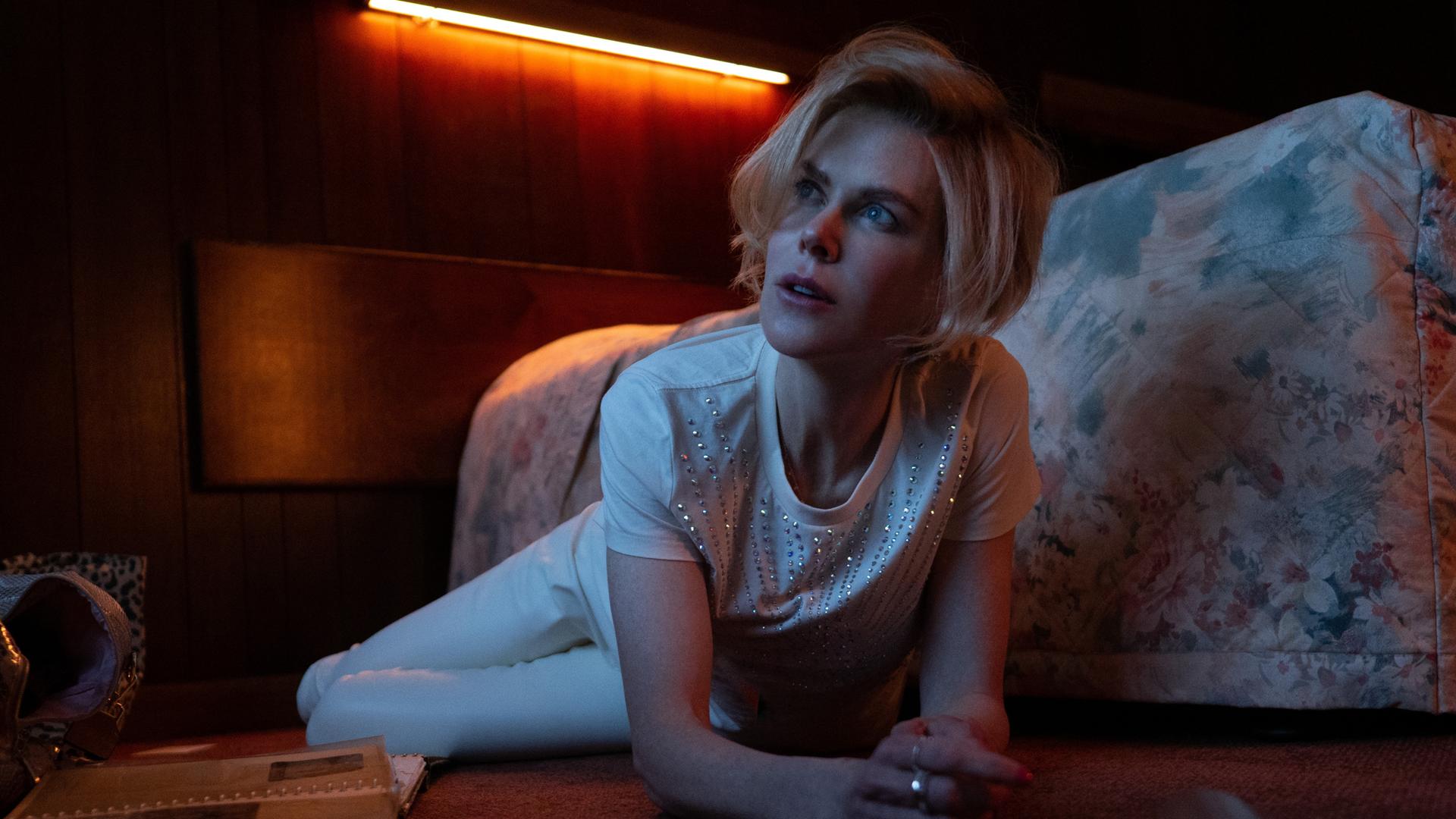 Die Schauspielerin Nicole Kidman liegt in einem Szenenfoto auf dem Teppichboden vor einem Bett mit geblümtem Überwurf. Sie trägt ein weißes Glitzershirt und weiße Hosen. Die Schauspielerin Nicole Kidman liegt in einem Szenenfoto auf dem Teppichboden vor einem Bett mit geblümtem Überwurf. Sie trägt ein weißes Glitzershirt und weiße Hosen.