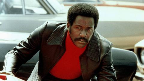 Der Schauspieler Richard Roundtree als Privatdetektiv John Shaft mit rotem Rollkragenpullover und schwarzer Lederjacke. Er beugt sich über die Motorhaube eines roten Autos.