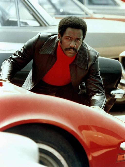 Der Schauspieler Richard Roundtree als Privatdetektiv John Shaft mit rotem Rollkragenpullover und schwarzer Lederjacke. Er beugt sich über die Motorhaube eines roten Autos.
