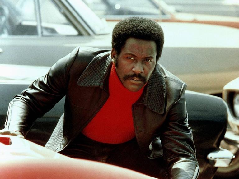 Der Schauspieler Richard Roundtree als Privatdetektiv John Shaft mit rotem Rollkragenpullover und schwarzer Lederjacke. Er beugt sich über die Motorhaube eines roten Autos.