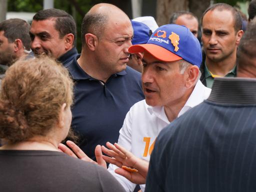 Nikol Paschinjan steht in einer Menschenmenge und spricht gestikulierend. Er trägt eine blaue-orangene Basecap.