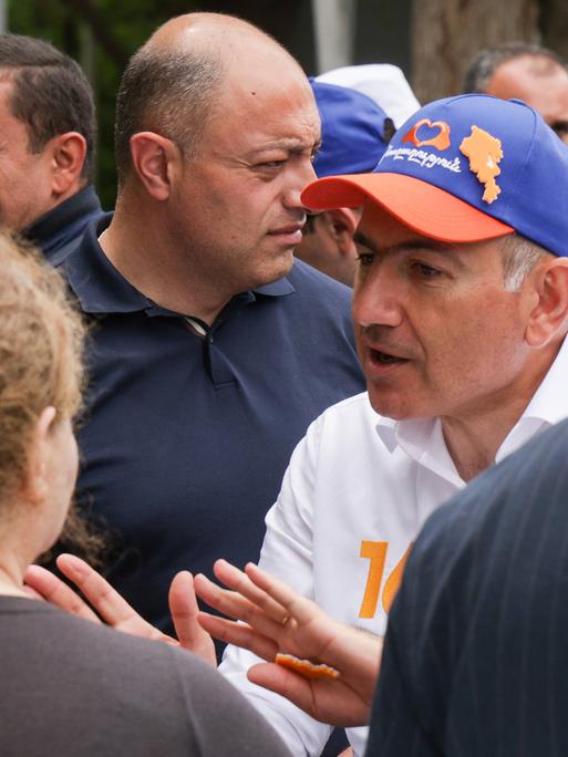 Nikol Paschinjan steht in einer Menschenmenge und spricht gestikulierend. Er trägt eine blaue-orangene Basecap.