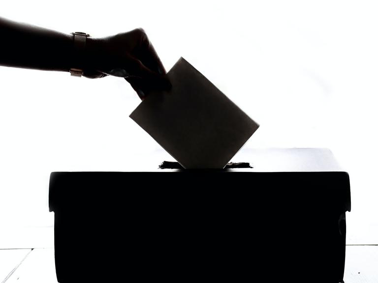 Eine Hand hält einen Wahlumschlag, kurz bevor er in einer Wahlurne gesteckt wird.