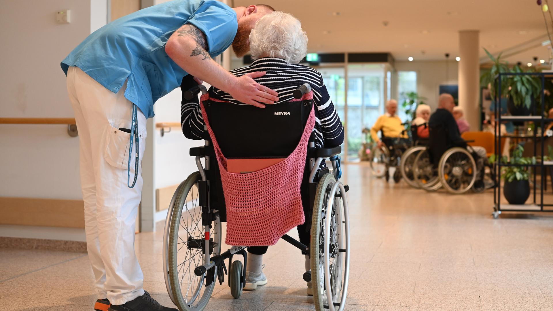 Ein Pfleger beugt sich hinunter zu einer Frau mit grauen Haaren, die man von hinten sieht und welche in einem Rollstuhl sitzt. Ein Pfleger beugt sich hinunter zu einer Frau mit grauen Haaren, die man von hinten sieht und welche in einem Rollstuhl sitzt.