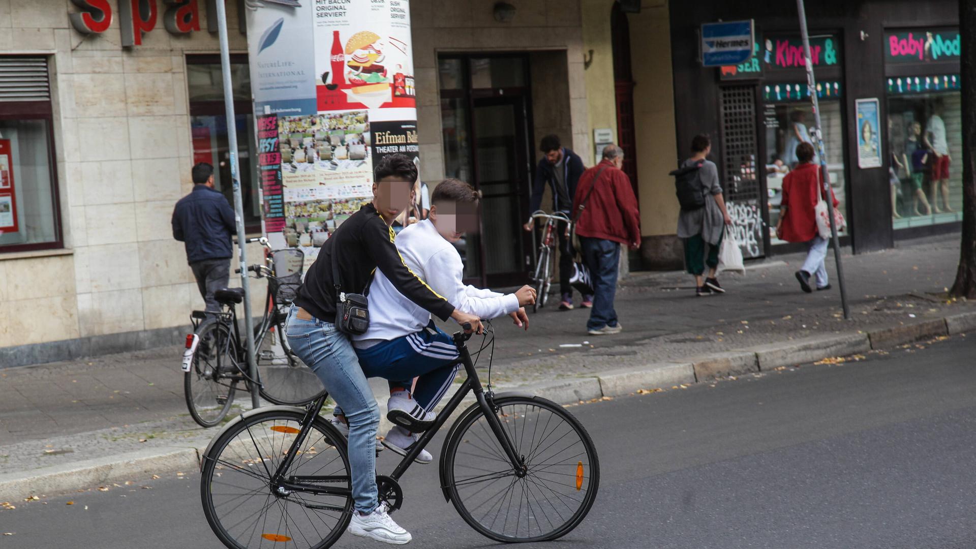 Zwei Jugendliche fahren mit einem Fahrrad – der eine auf der Stange des Fahrrads – auf der Karl-Marx-Allee in Neukölln und behindern den Verkehr.