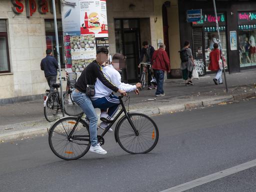 Zwei Jugendliche fahren mit einem Fahrrad – der eine auf der Stange des Fahrrads – auf der Karl-Marx-Allee in Neukölln und behindern den Verkehr.