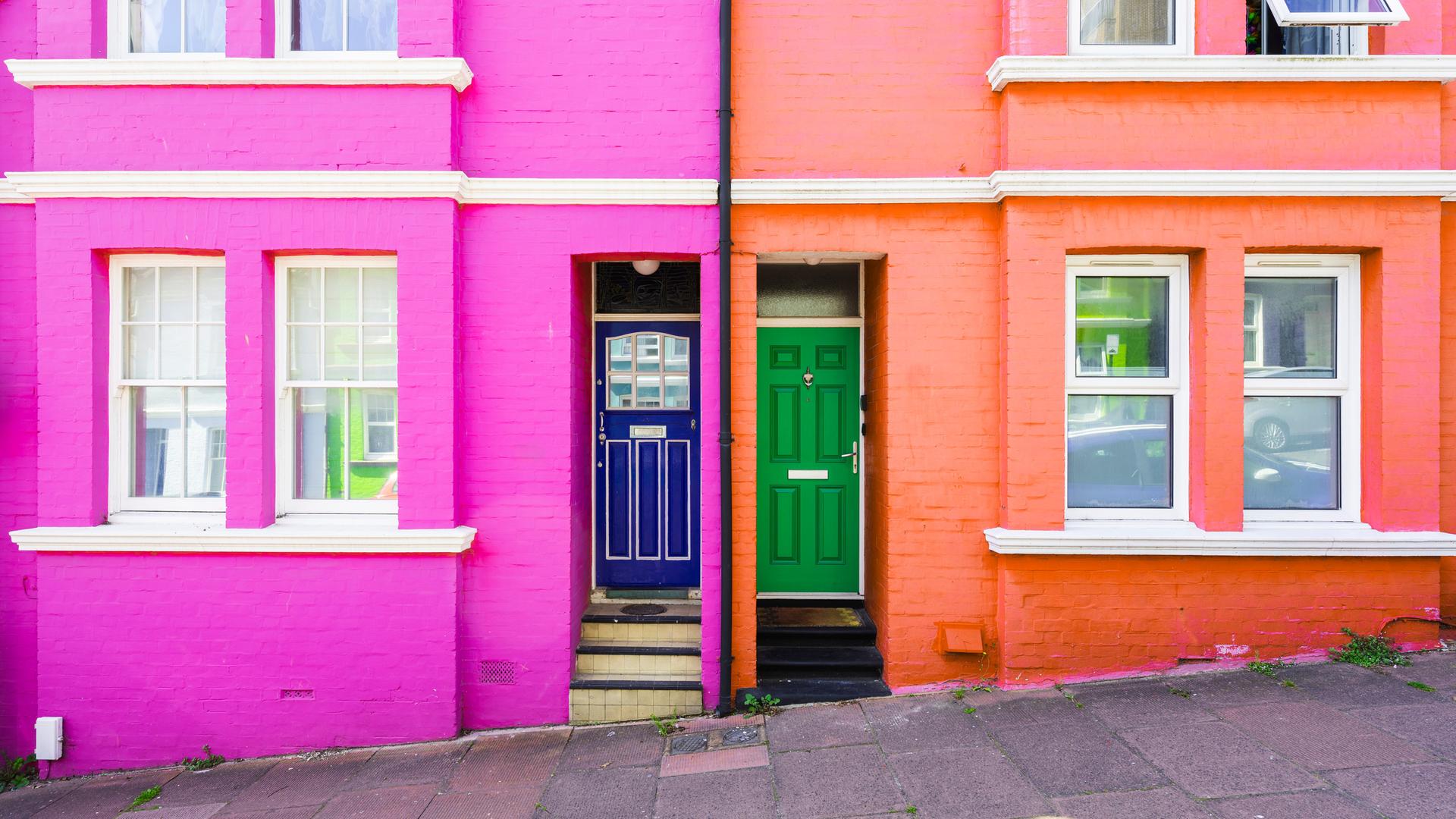 Zwei farbig gestrichene Häuser nebeneinander. Das Haus mit pinker Fassade hat eine blaue Tür, das Haus mit oranger Fassade eine grüne Tür. Zwei farbig gestrichene Häuser nebeneinander. Das Haus mit pinker Fassade hat eine blaue Tür, das Haus mit oranger Fassade eine grüne Tür.