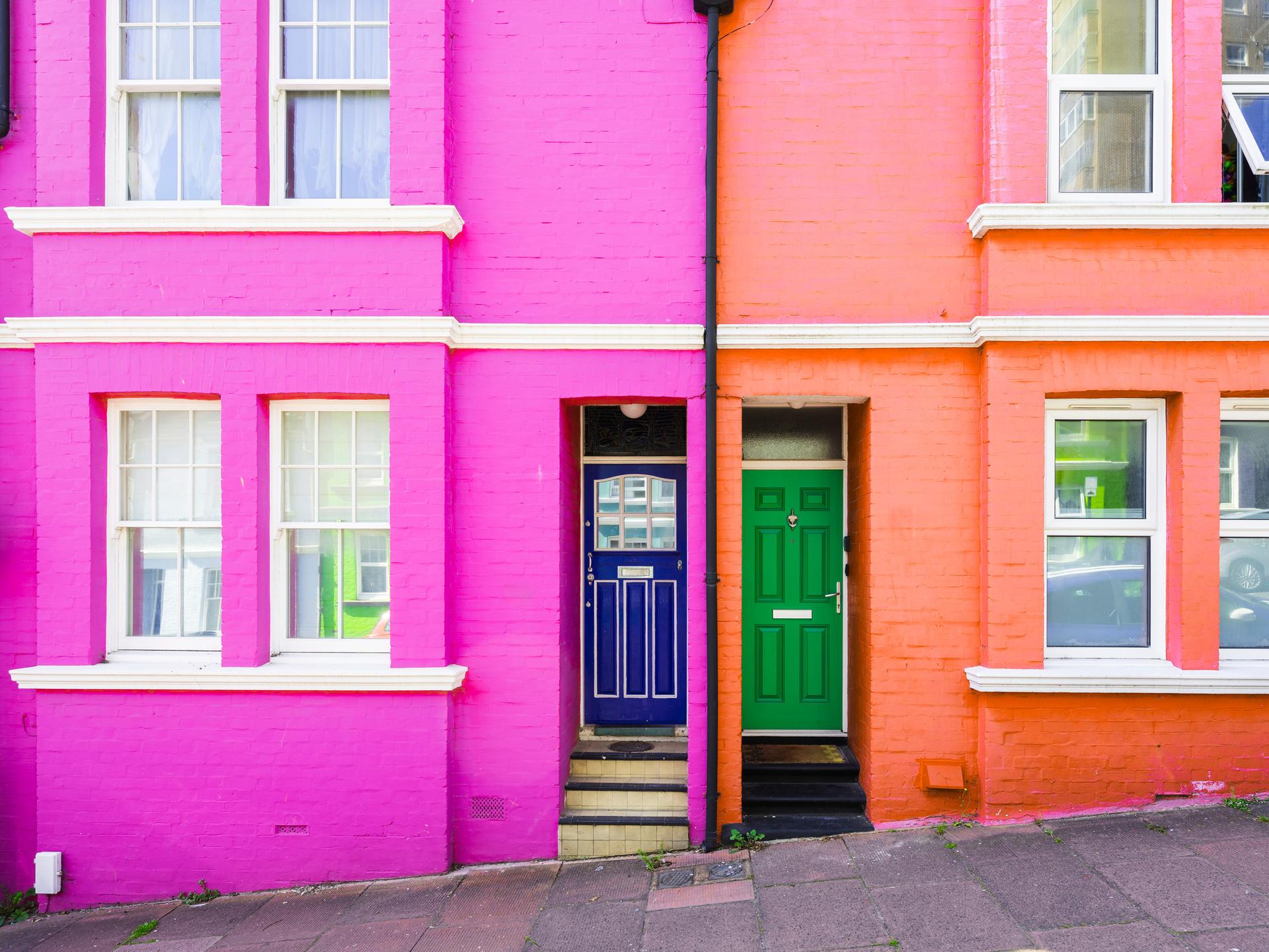 Zwei farbig gestrichene Häuser nebeneinander. Das Haus mit pinker Fassade hat eine blaue Tür, das Haus mit oranger Fassade eine grüne Tür.