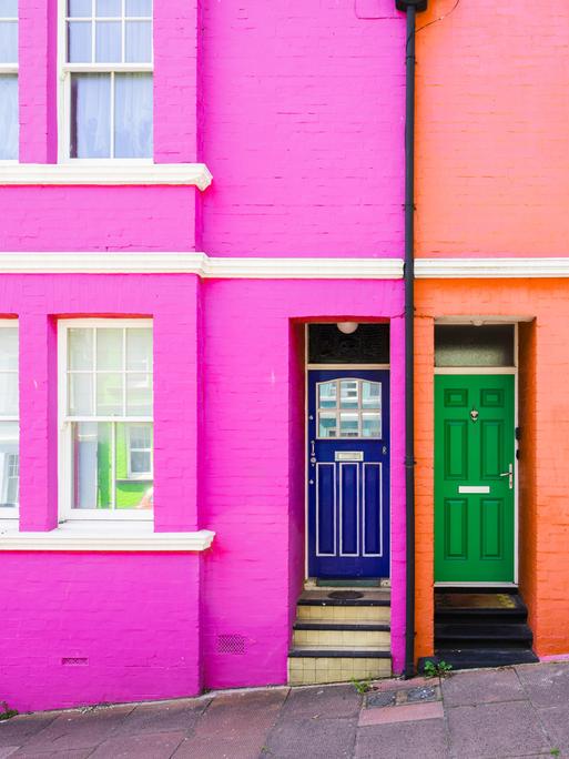 Zwei farbig gestrichene Häuser nebeneinander. Das Haus mit pinker Fassade hat eine blaue Tür, das Haus mit oranger Fassade eine grüne Tür.