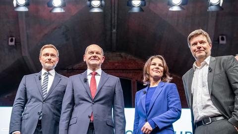 Christian Lindner,  Olaf Scholz, Annalena Baerbock und Robert Habeck stehen bei einer Pressekonferenz auf einer Bühne. Im Hintergrund sind Scheinwerfer zu sehen.