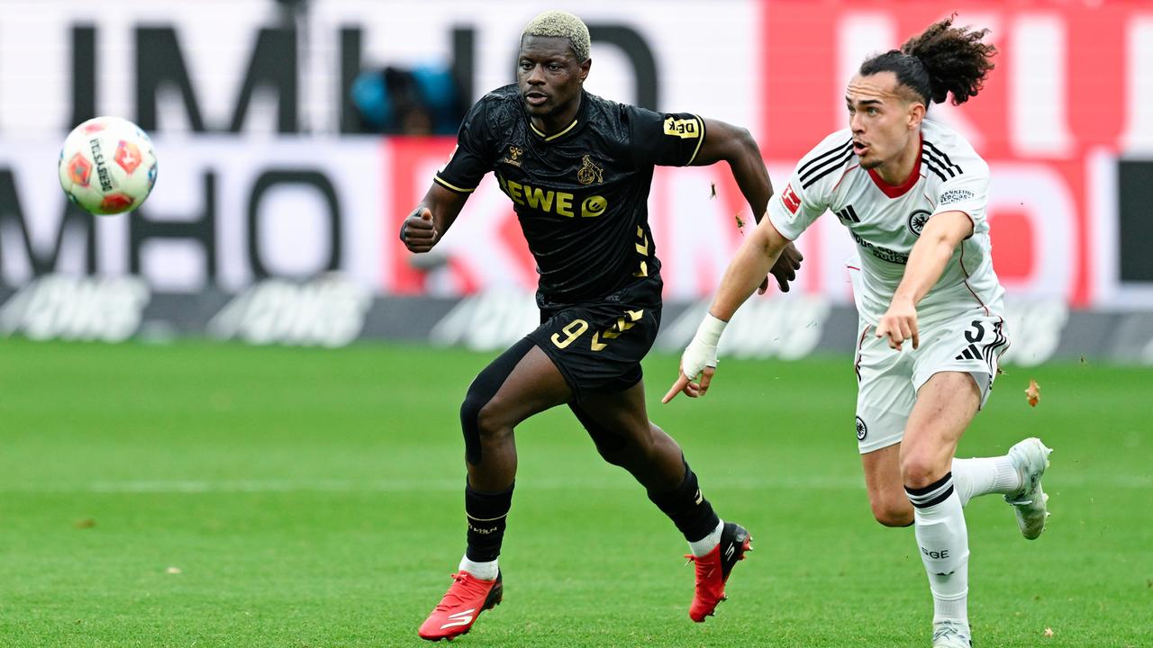 Das Bild zeigt zwei Fußball-Profis auf einem Rasenplatz im vollen Lauf. Beide versuchen als erster an den Fußball zu kommen. Im Hintergrund ist schemenhaft eine Bandenwerbung am Spielfeldrand zu sehen.