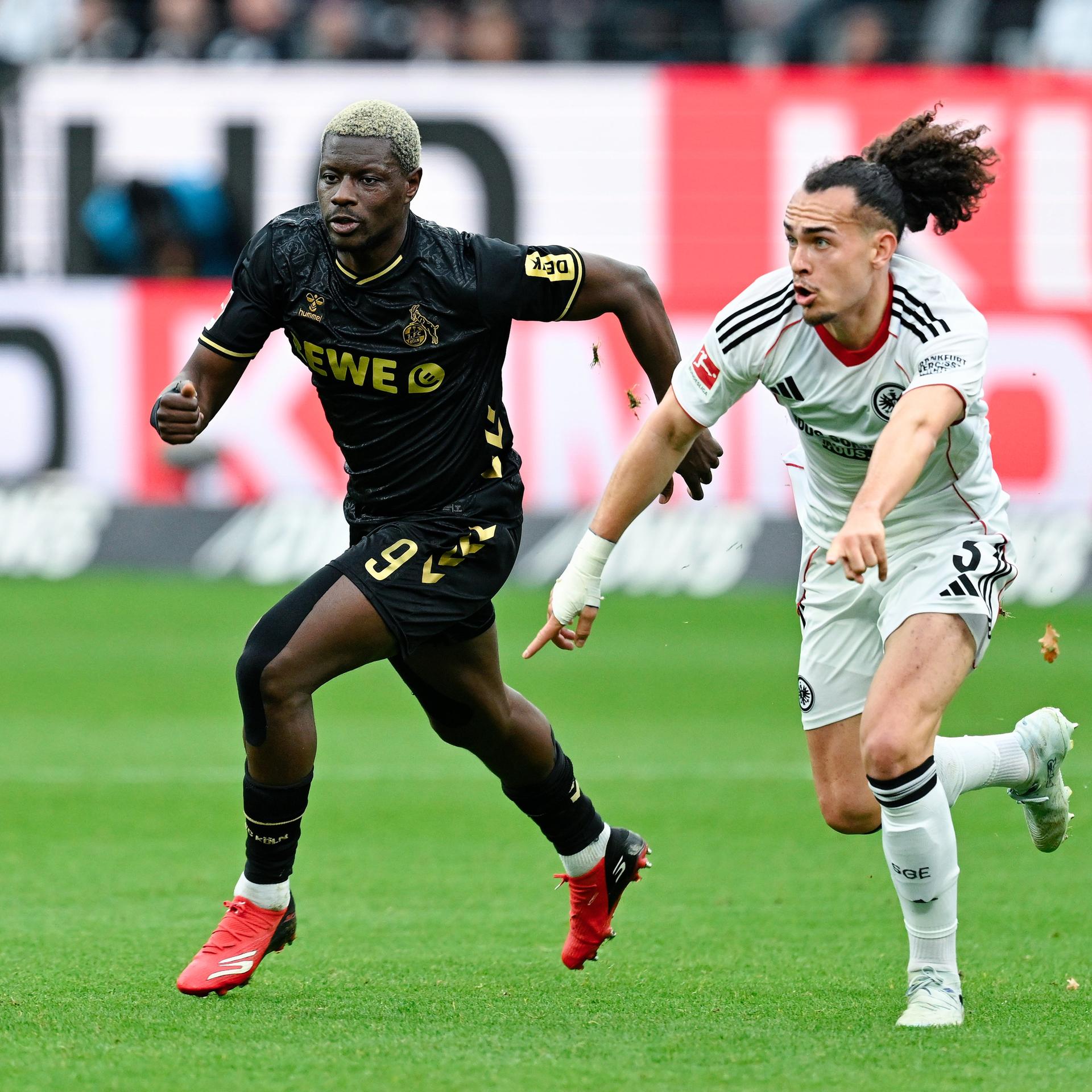 Das Bild zeigt zwei Fußball-Profis auf einem Rasenplatz im vollen Lauf. Beide versuchen als erster an den Fußball zu kommen. Im Hintergrund ist schemenhaft eine Bandenwerbung am Spielfeldrand zu sehen.