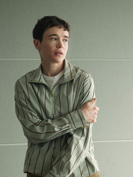 Elliot Page steht mit leicht verschränkten Armen, zur Seite schauend vor einer hellen Wand und trägt ein gestreiftes Hemd.