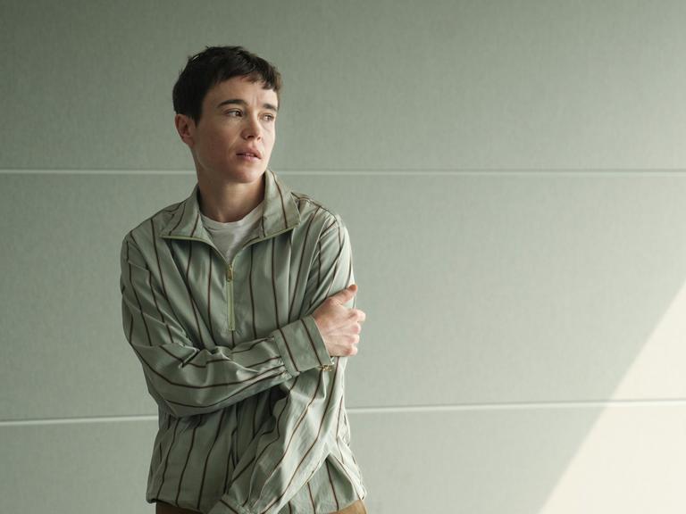 Elliot Page steht mit leicht verschränkten Armen, zur Seite schauend vor einer hellen Wand und trägt ein gestreiftes Hemd.