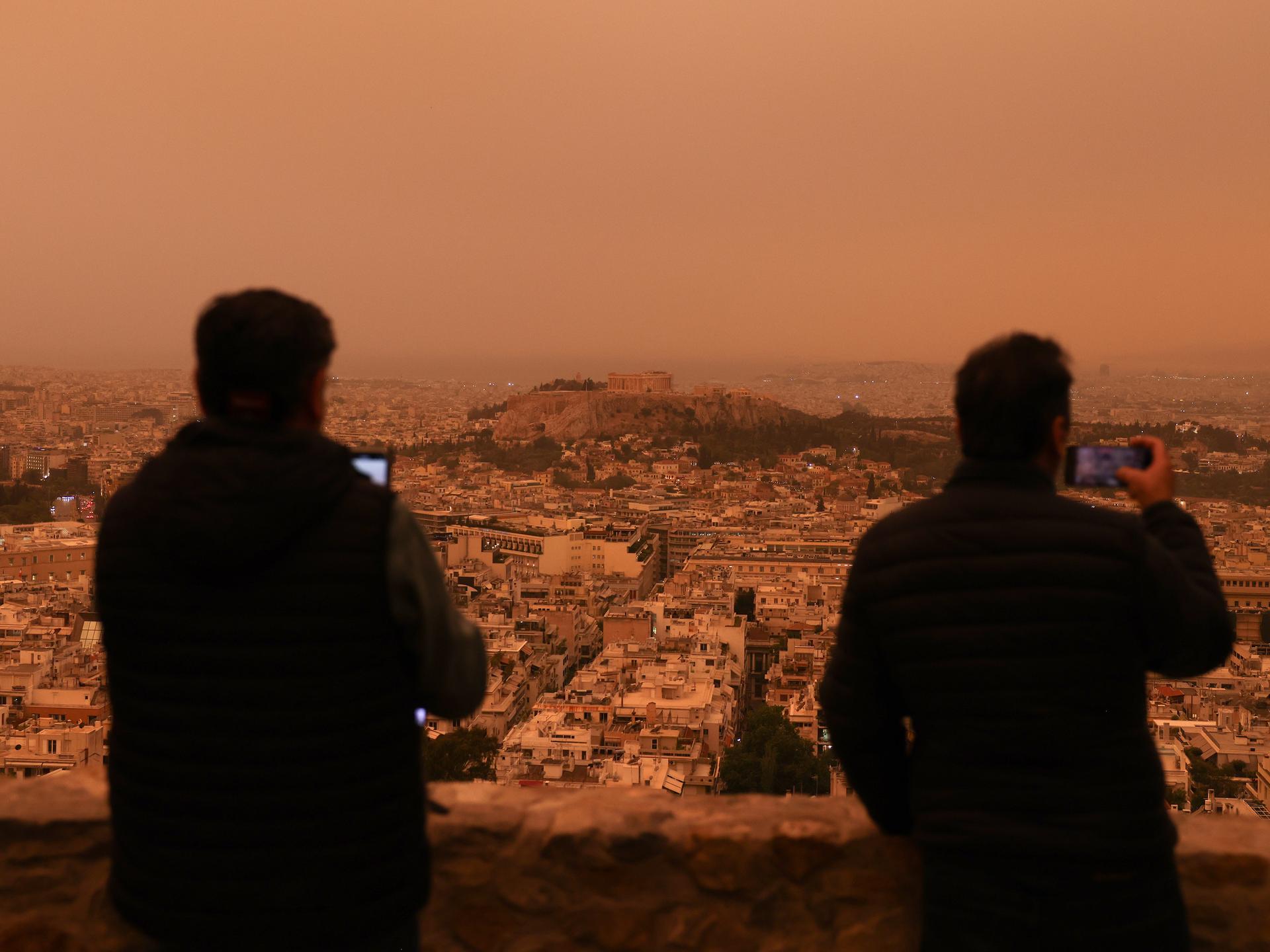 Staub aus der Wüstse hat sich über die griechische Hauptstadt Athen gelegt. Zwei Menschen sitzen auf einer Anhöhe und blicken auf die Stadt, die rot-gelblisch schimmert.