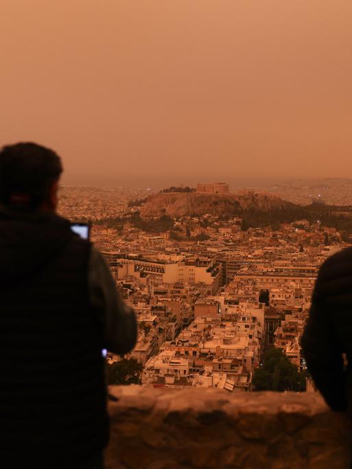 Staub aus der Wüstse hat sich über die griechische Hauptstadt Athen gelegt. Zwei Menschen sitzen auf einer Anhöhe und blicken auf die Stadt, die rot-gelblisch schimmert.