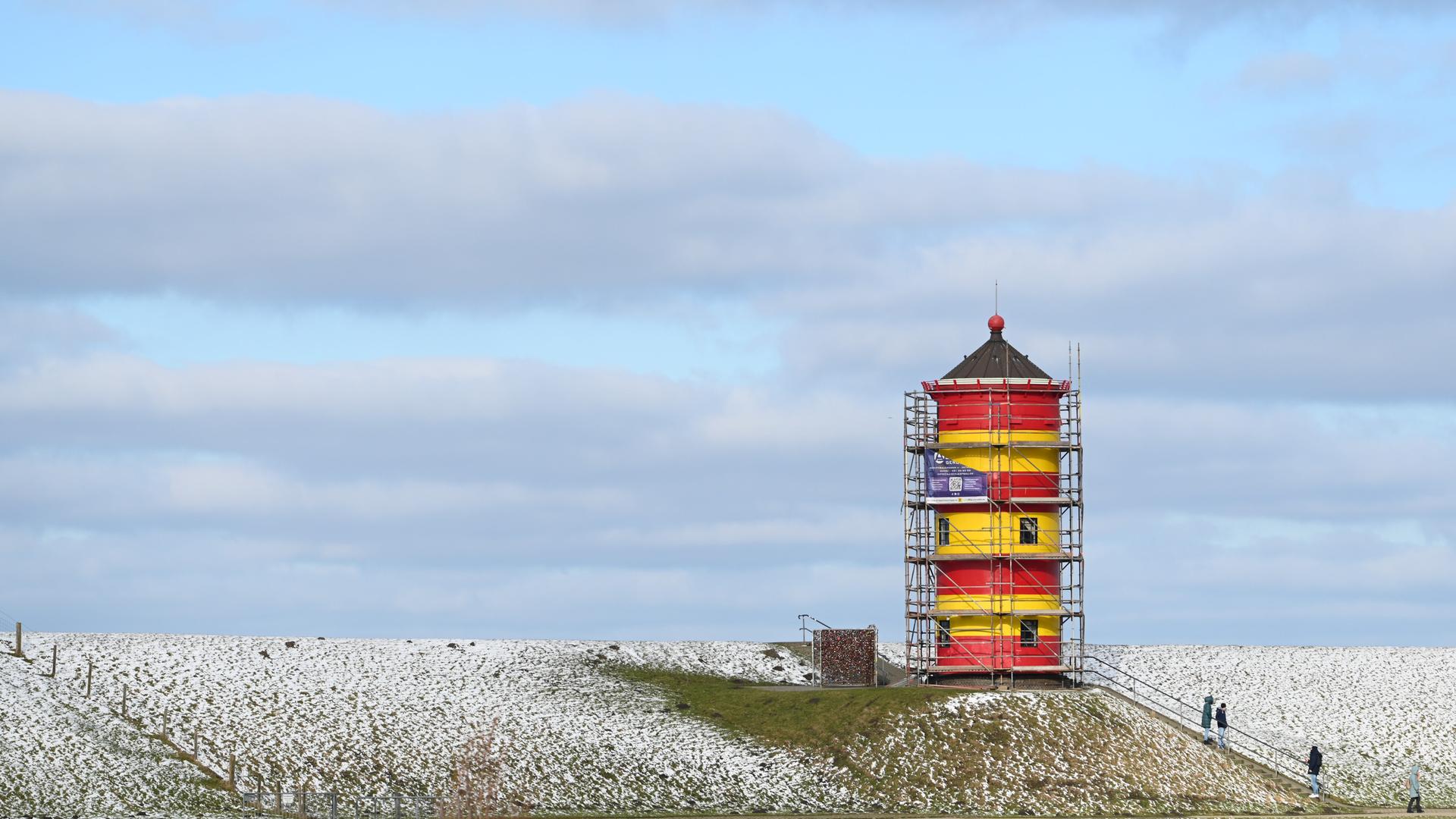 Der Leuchtturm von Pilsum im Winter. Der Turm ist ziemlich klein und rot-gelb angestrichen. Rund um den Turm ist ein Gerüst. 