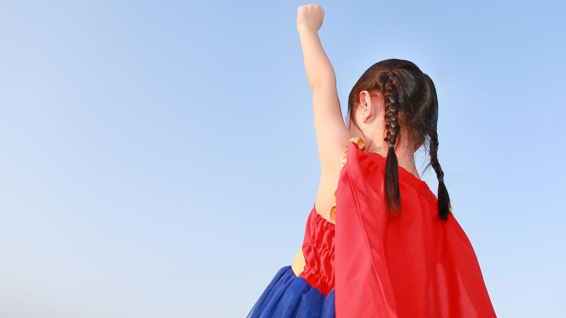 Ein Mädchen im Kindergartenalter trägt ein Super(wo)man-Kostüm und streckt eine Faust nach oben.