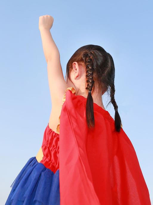 Ein Mädchen im Kindergartenalter trägt ein Super(wo)man-Kostüm und streckt eine Faust nach oben.