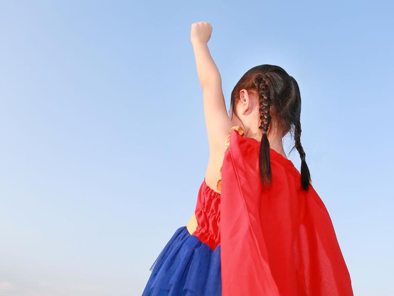 Ein Mädchen im Kindergartenalter trägt ein Super(wo)man-Kostüm und streckt eine Faust nach oben.