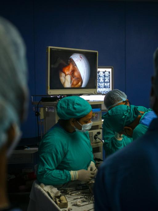 Ein Team aus Ärztinnen und Ärzten behandelt einen Patienten mit Mukormykose-Infektion während der Coronapandemie. Auf einem Bildschirm im Hintergrund ist das Gesicht des Patienten zu sehen.