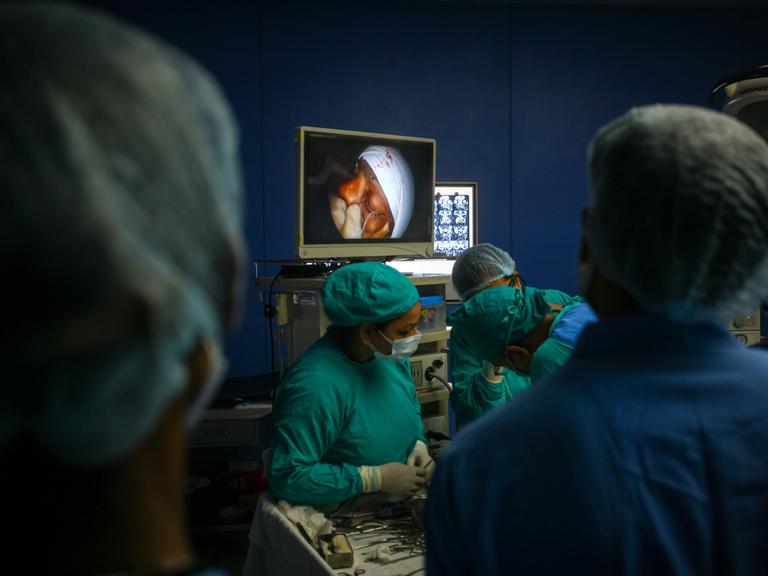 Ein Team aus Ärztinnen und Ärzten behandelt einen Patienten mit Mukormykose-Infektion während der Coronapandemie. Auf einem Bildschirm im Hintergrund ist das Gesicht des Patienten zu sehen.