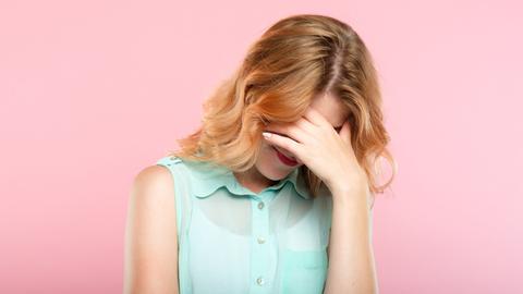 Eine Frau in mintgrüner Bluse vor pinkem Hintergrund, hält sich vor Scham die Hand vor das Gesicht.