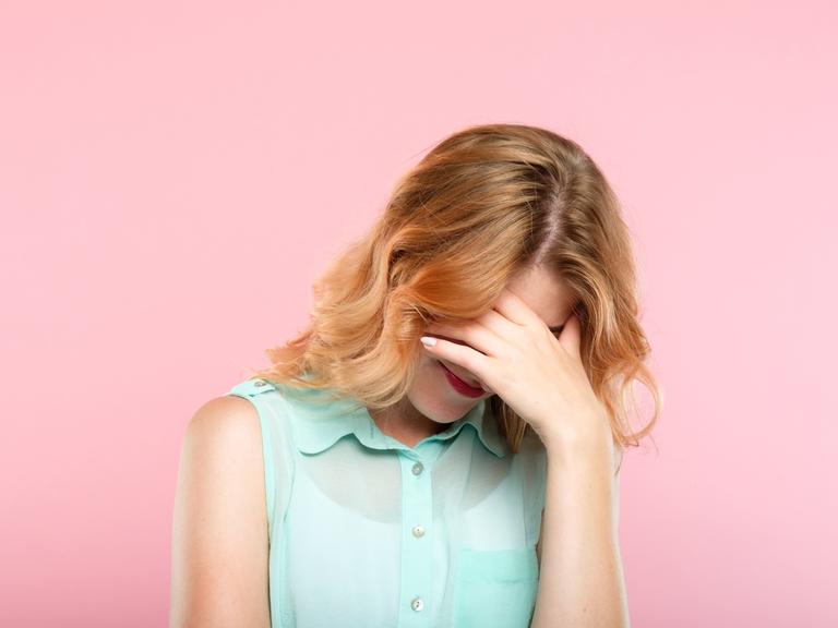 Eine Frau in mintgrüner Bluse vor pinkem Hintergrund, hält sich vor Scham die Hand vor das Gesicht.