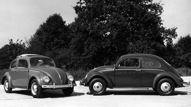 Schwarzweißes Archivfoto zweier VW Käfers Export-Modelle auf einem Weg. Im Hintergrund sind Bäume sichtbar.