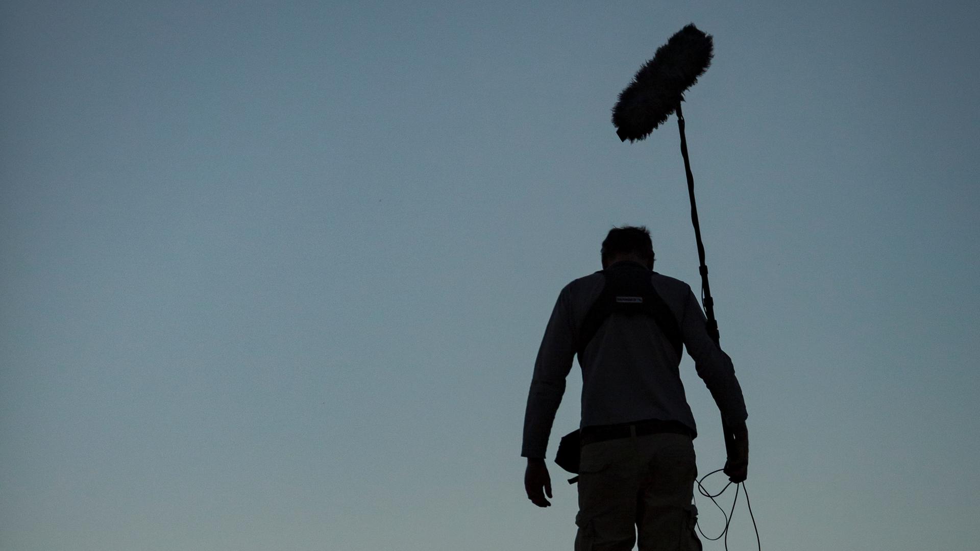 Ein Toningenieur einer Dokumentarfilmcrew geht eine Straße entlang, während im Hintergrund der Himmel dunkler wird.