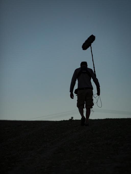 Ein Toningenieur einer Dokumentarfilmcrew geht eine Straße entlang, während im Hintergrund der Himmel dunkler wird. Ein Toningenieur einer Dokumentarfilmcrew geht eine Straße entlang, während im Hintergrund der Himmel dunkler wird.