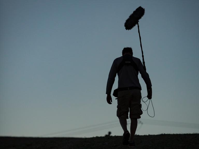 Ein Toningenieur einer Dokumentarfilmcrew geht eine Straße entlang, während im Hintergrund der Himmel dunkler wird.