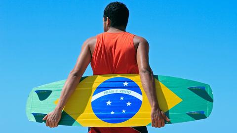 Ein Mann trägt ein Kitesurfing-Board, das als brasilianische Flagge bemalt ist.