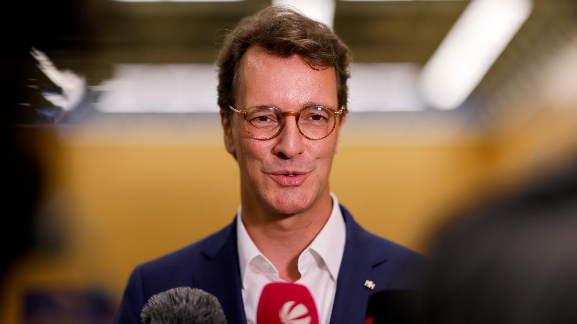 Hendrik Wüst spricht für ein Interview in Mikrofone. Er ist ein mittelalter Mann im dunkelblauen Anzug und mit einer braunen runden Hornbrille.