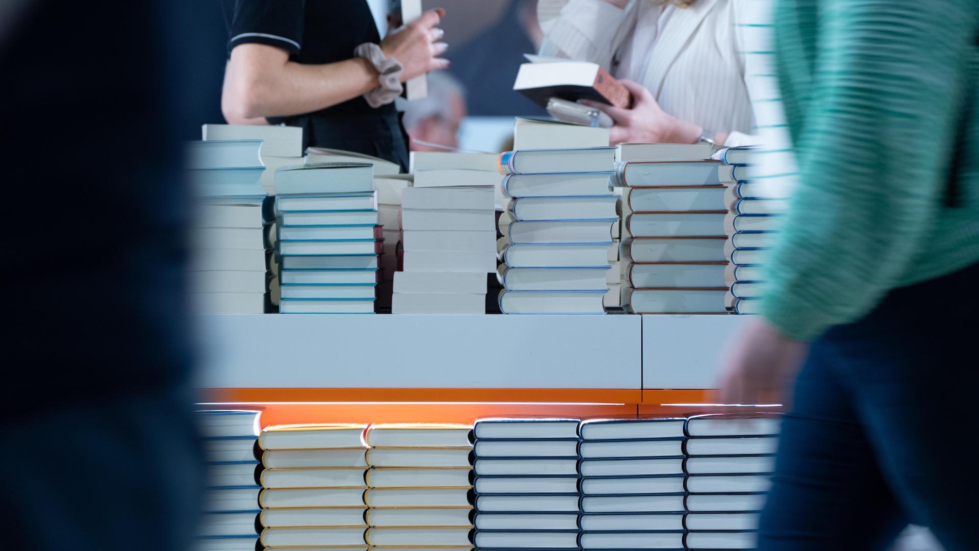 Bücherstapel sind am Stand eines Verlages aufgetürmt. Besucherinnen halten Bücher in der Hand. Der Verlag präsentiert sich in einer Halle der Frankfurter Buchmesse (2024).