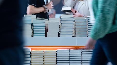 Bücherstapel sind am Stand eines Verlages aufgetürmt. Besucherinnen halten Bücher in der Hand. Der Verlag präsentiert sich in einer Halle der Frankfurter Buchmesse (2024).