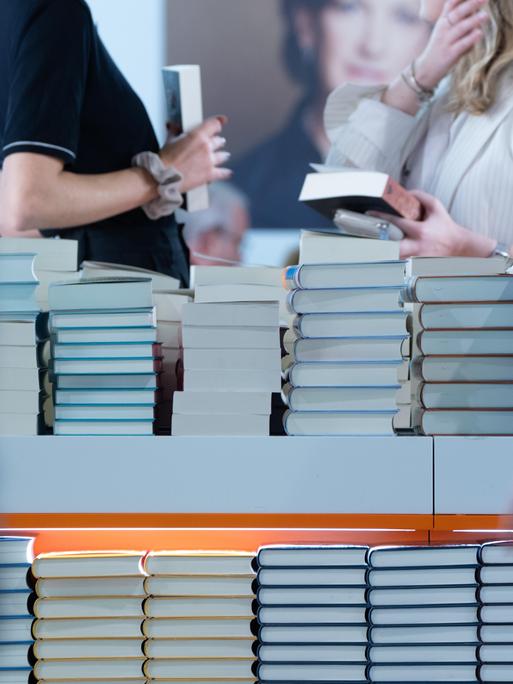 Bücherstapel sind am Stand eines Verlages aufgetürmt. Besucherinnen halten Bücher in der Hand. Der Verlag präsentiert sich in einer Halle der Frankfurter Buchmesse (2024).