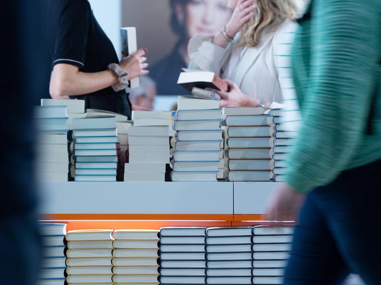 Bücherstapel sind am Stand eines Verlages aufgetürmt. Besucherinnen halten Bücher in der Hand. Der Verlag präsentiert sich in einer Halle der Frankfurter Buchmesse (2024).